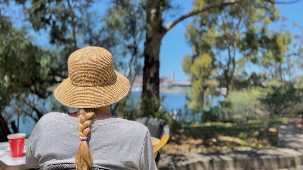 Woman sitting outdoors by the water wearing a straw hat, relaxing in nature and enjoying a peaceful mindful moment.